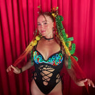 A woman posing in front of a red curtain backdrop, wearing a black strappy bodysuit with a vibrant multicolor neon-style graphic across the chest, yellow and green hair extensions, and red sheer fabric draped over her arms.